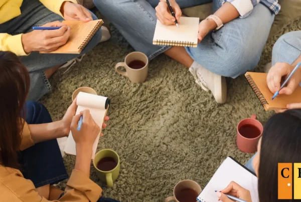 Small peer support group sitting in a circle on the floor with notebooks and mugs of tea, writing and connecting together in a warm informal setting
