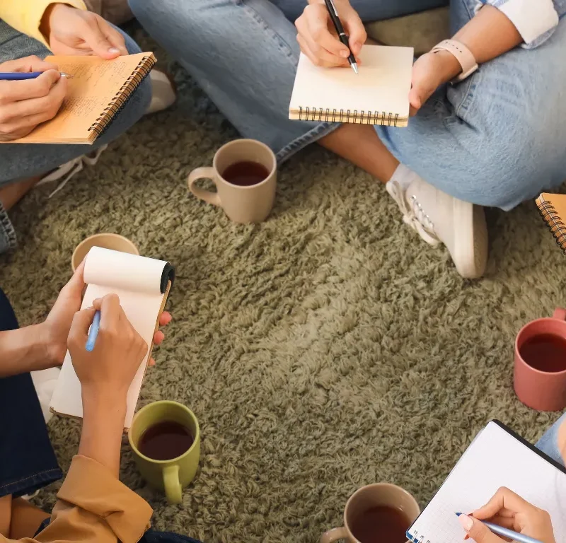 Small peer support group sitting in a circle on the floor with notebooks and mugs of tea, writing and connecting together in a warm informal setting